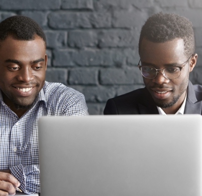 Two happy African colleagues using laptop. Manager in shirt showing presentation on notebook computer to his boss wearing suit and glasses, during meeting in modern office interior with brick walls