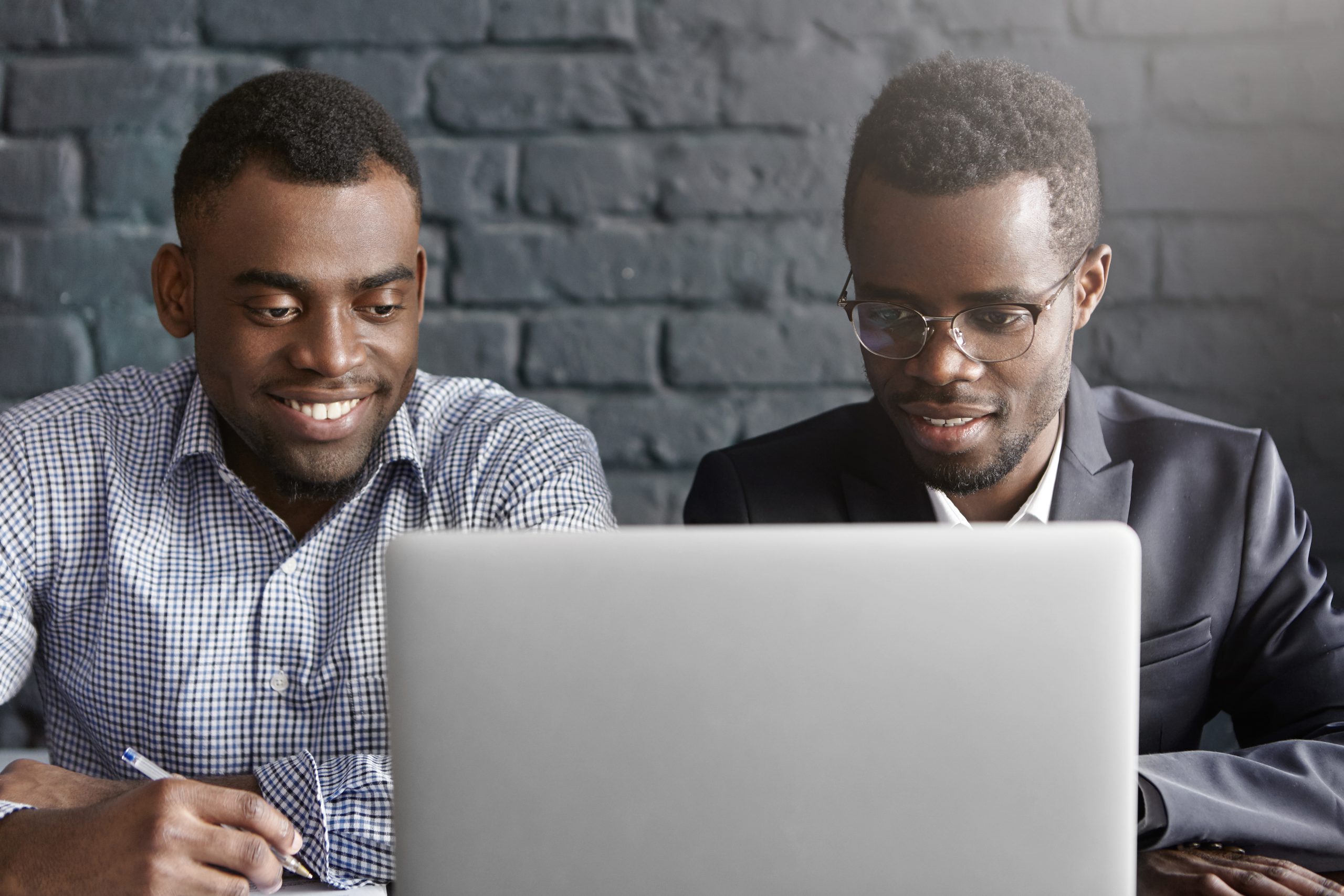 Two happy African colleagues using laptop. Manager in shirt showing presentation on notebook computer to his boss wearing suit and glasses, during meeting in modern office interior with brick walls